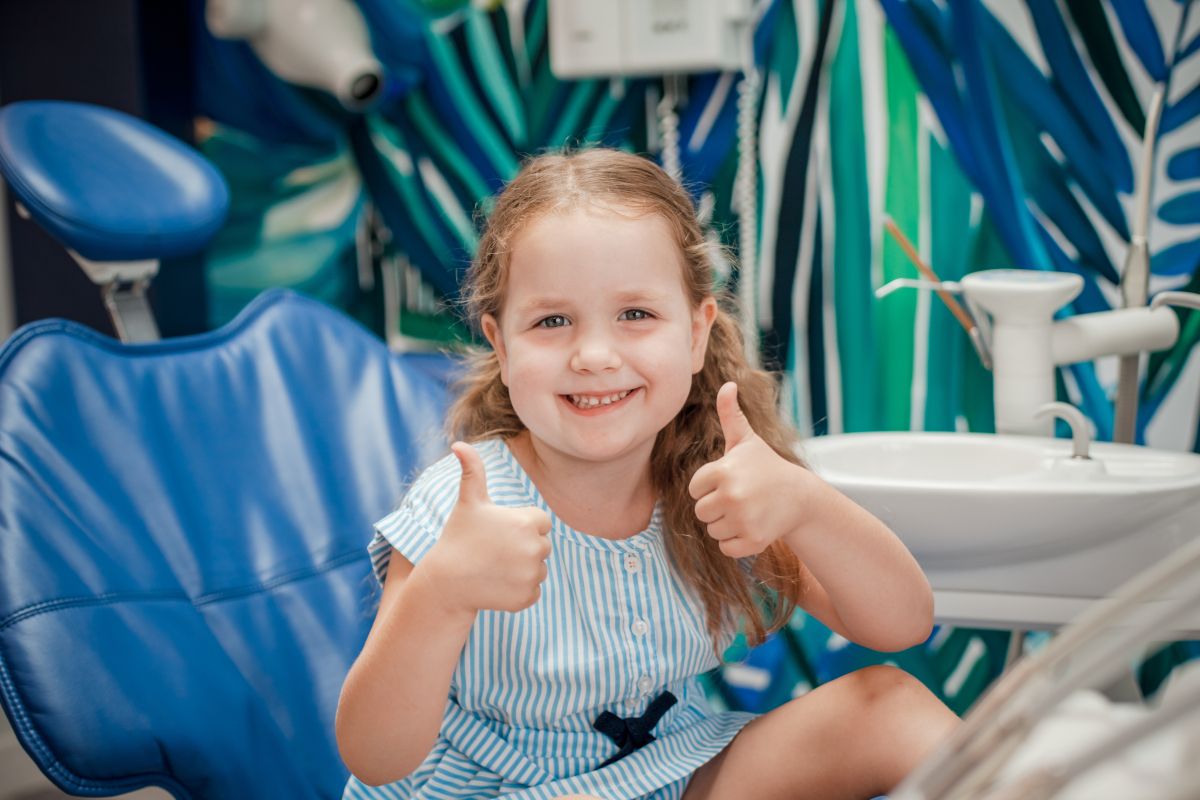 Young girl giving thumbs up gesture, smiling at camera with blue tropical leaf backdrop.