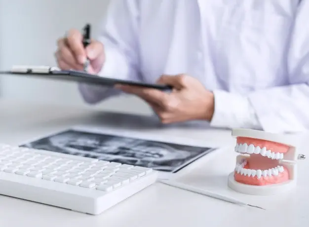Dentist reviewing dental X-ray and treatment plan with teeth model on desk.
