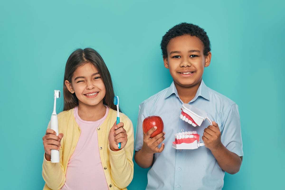 Two smiling children with healthy teeth holding toothbrushes and an apple, promoting pediatric dental care in McKinney.